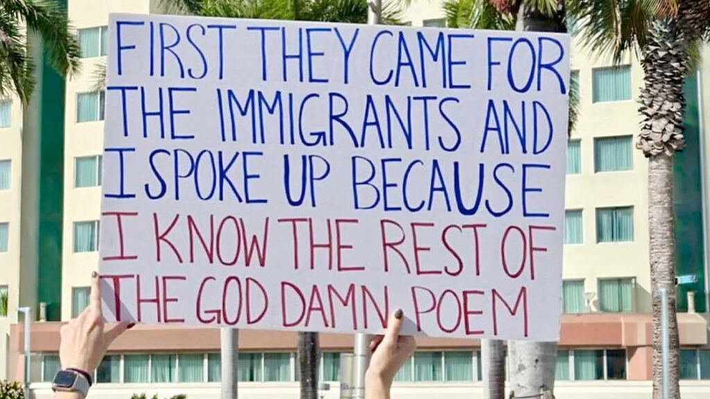 Person holds a large poster with blue and red text reading: 'First they came for the immigrants and I spoke up because I know the rest of the god dam n poem' outdoors with buildings and palm trees in the background,