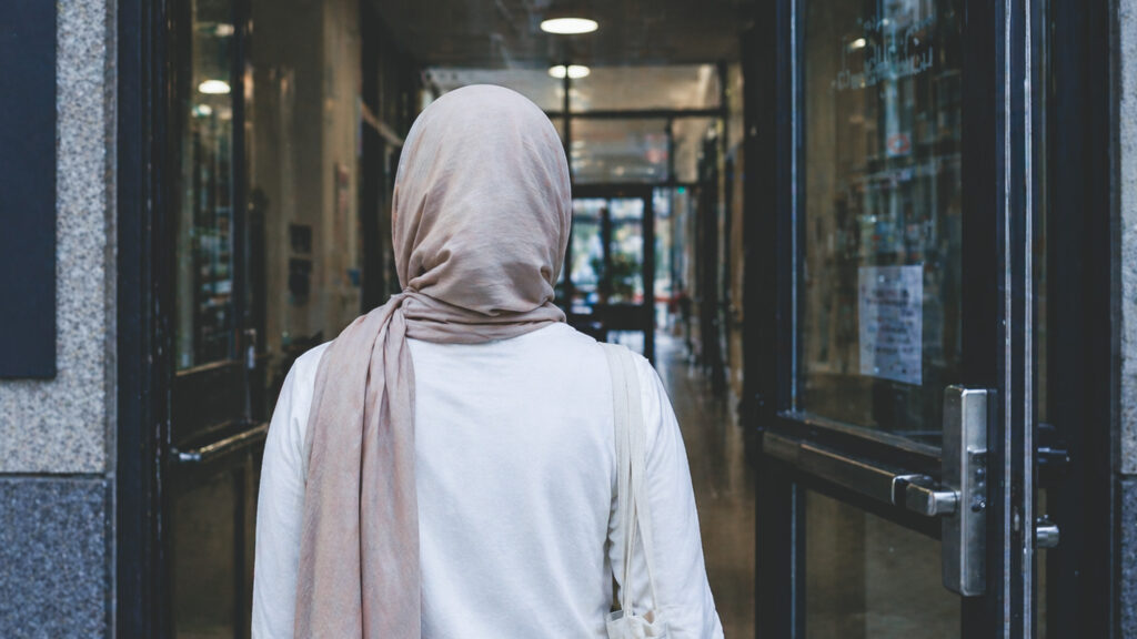 Back view of a person wearing a beige hijab and white top standing in a glass-front doorway in an urban building corridor.