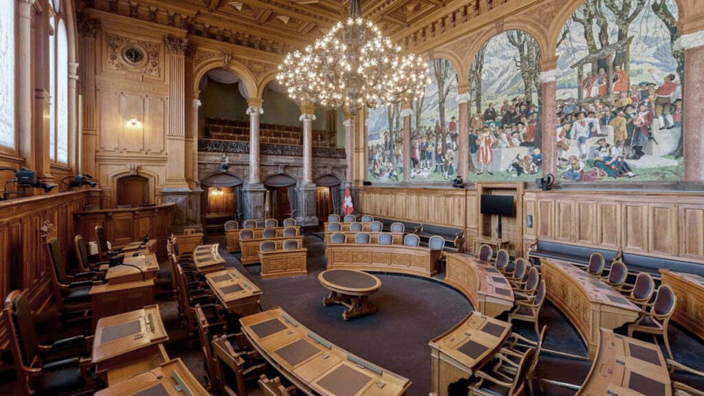 Ornate parliamentary chamber with a central round dais, surrounding wooden desks, and a grand chandelier; mural wall depicts a historical scene.