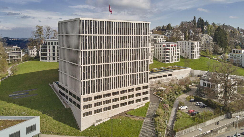 Aerial view of a tall white office building with grid-like windows on a grassy hill, a Swiss flag atop the roof.